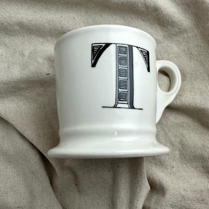 May include: White ceramic mug with a black letter "T" design. The mug has a rounded handle and a flared base. The letter "T" is stylized with a decorative pattern. The mug is on a neutral-colored fabric.