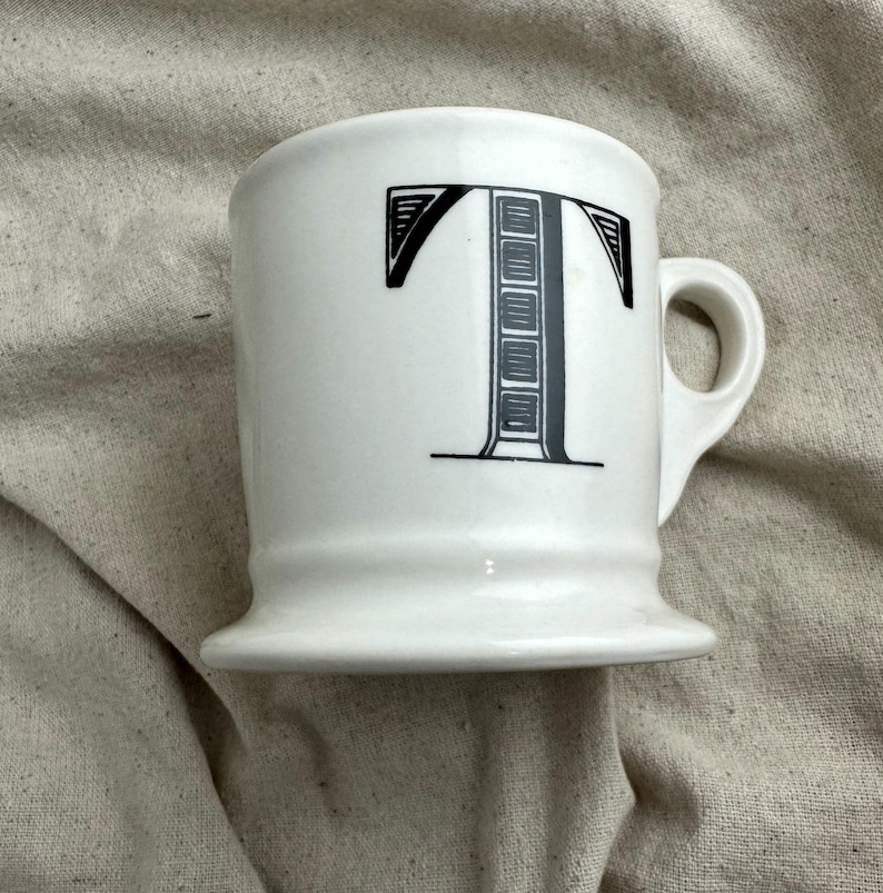 May include: White ceramic mug with a black letter "T" design. The mug has a rounded handle and a flared base. The letter "T" is stylized with a decorative pattern. The mug is on a neutral-colored fabric.