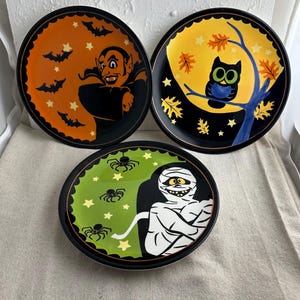 May include: Three Halloween-themed ceramic plates with black rims. The first plate features a cartoon vampire with orange and black bats and stars on a black background. The second plate features a cartoon owl perched on a branch with yellow and orange leaves on a black background. The third plate features a cartoon mummy with green and yellow stars and spiders on a black background.