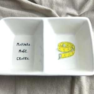 May include: White rectangular divided dish with the words "Measure. Make. Create." in black script on one side. The other side features a yellow tape measure illustration. The dish is on a neutral-colored fabric.