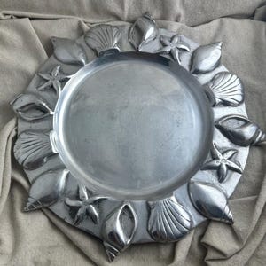 May include: A silver-colored decorative tray with a circular center and a border adorned with various seashell and starfish designs. The tray is made of metal and has a polished finish, perfect for coastal decor.