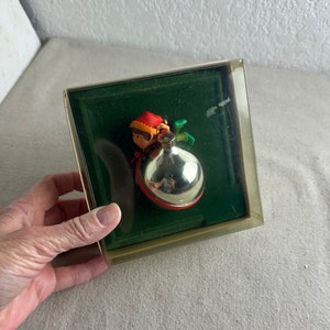 May include: A vintage Christmas bauble featuring a small elf figurine holding a silver bauble. The ornament is displayed in a clear plastic box with a green velvet lining.