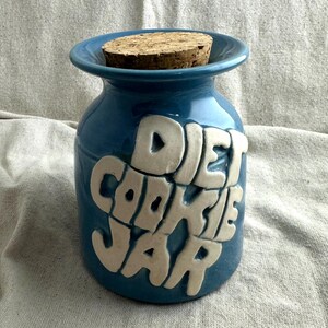 May include: A blue ceramic cookie jar with the words "DIET COOKIE JAR" in white raised letters. The jar has a cork stopper and a wide rim. The jar is on a neutral background.