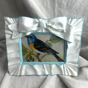 May include: A white picture frame with a blue bird illustration. The frame has a ruffled edge and a bow with blue polka dots. The frame's inner border is light blue.