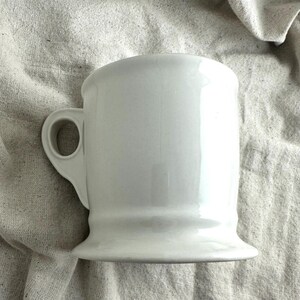 May include: A white ceramic mug with a rounded top and a sturdy handle. The mug has a flared base and a smooth, glossy finish. The mug is designed for beverages.