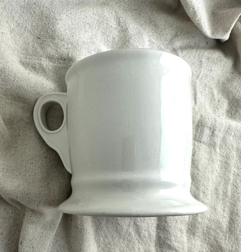 May include: A white ceramic mug with a rounded top and a sturdy handle. The mug has a flared base and a smooth, glossy finish. The mug is designed for beverages.