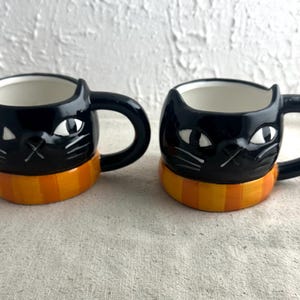 May include: Two black ceramic cat-shaped mugs with white eyes and whiskers. Each mug has a handle and an orange and yellow striped band around the base. The mugs are on a neutral-colored surface.