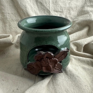 May include: A handcrafted, green ceramic oil warmer with a dark brown magnolia flower accent. The oil warmer has a round shape with a wide opening and a decorative flower on the front.