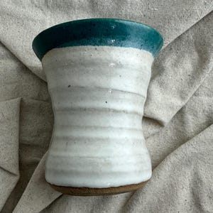 May include: A handmade ceramic vase with a white and teal glaze. The vase has a unique, curved shape and a textured surface. The top rim is teal, transitioning to white. The base is a natural, unglazed brown. A decorative piece for the home.