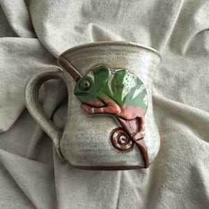 May include: A ceramic mug with a speckled off-white glaze featuring a raised, three-dimensional green and orange chameleon sculpture wrapped around the side. The mug has a curved handle and a wide rim, showcasing a handcrafted pottery aesthetic.