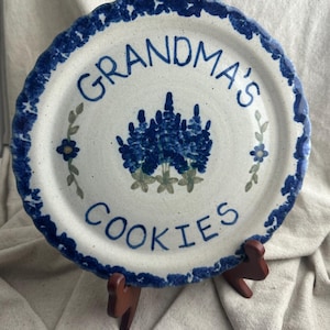 May include: A white ceramic plate with a blue floral design and the words "GRANDMA'S COOKIES" in blue script. The plate has a scalloped edge with a blue border and is supported by a wooden stand.