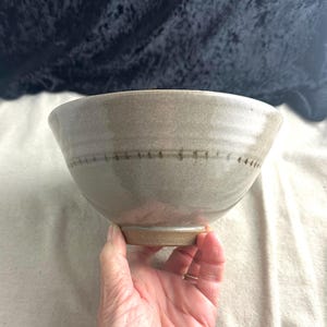 May include: A light gray ceramic bowl with a brown line around the rim. The bowl has a slightly textured surface and a rounded bottom.