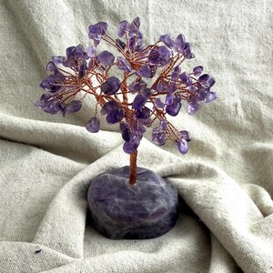 May include: A decorative amethyst tree sculpture. The tree has a copper wire frame with small amethyst gemstone leaves and a gemstone base. The amethyst gemstones are a deep purple colour. The tree is displayed on a neutral-coloured fabric.
