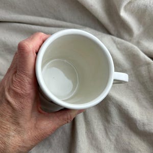 May include: A white ceramic mug with a handle, held in a hand. The interior of the mug is a creamy white color. The mug is set against a neutral-toned fabric background.