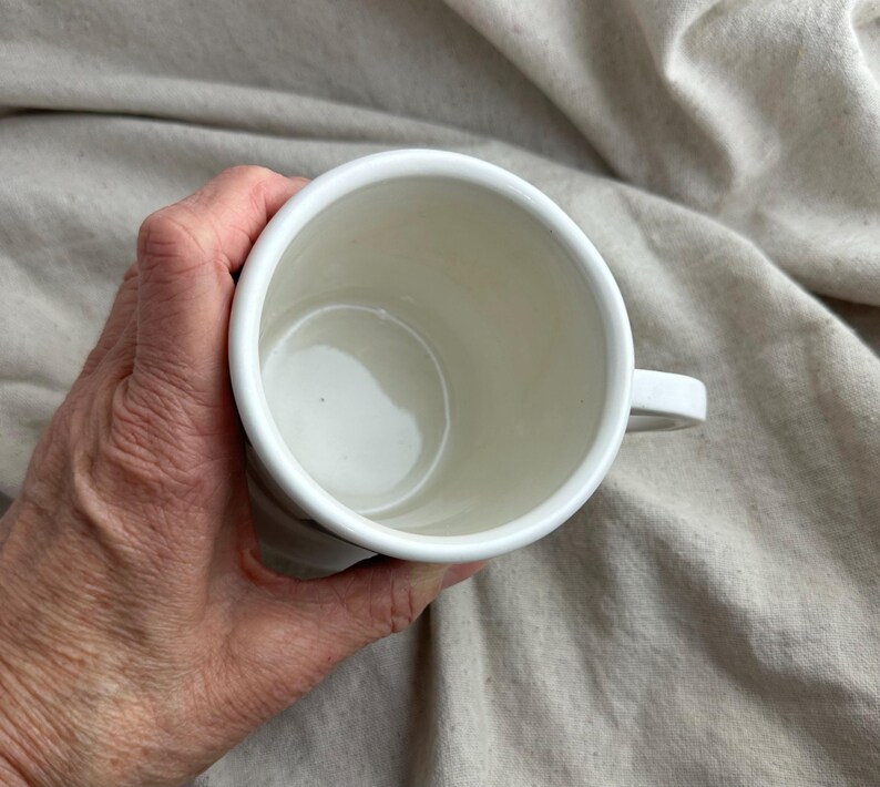 May include: A white ceramic mug with a handle, held in a hand. The interior of the mug is a creamy white color. The mug is set against a neutral-toned fabric background.