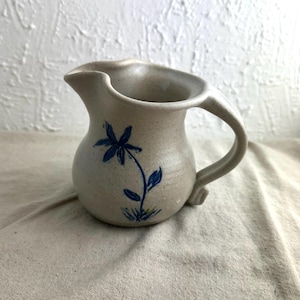 May include: A small, gray ceramic pitcher with a blue floral design. The pitcher has a rounded body and a handle.