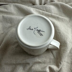 Op de afbeelding: Witte keramische koffiemok met een gebogen oor. De onderkant van de mok heeft de handtekening "Starla M. Hoffman" en de tekst "MADE IN CHINA". De mok staat op een beige stoffen ondergrond.