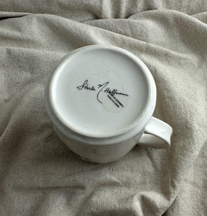 Op de afbeelding: Witte keramische koffiemok met een gebogen oor. De onderkant van de mok heeft de handtekening "Starla M. Hoffman" en de tekst "MADE IN CHINA". De mok staat op een beige stoffen ondergrond.