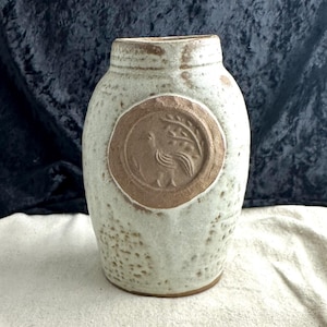 May include: A white ceramic vase with a brown circular design featuring a bird and a branch. The vase has a textured surface.