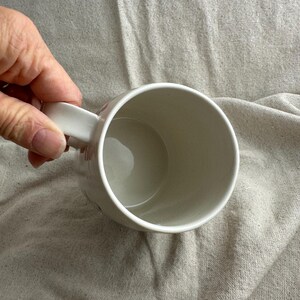 Op de afbeelding: Een witte keramische mok met een gebogen handvat. De mok is leeg en heeft een gladde, glanzende afwerking. De binnenkant van de mok is lichtbeige.