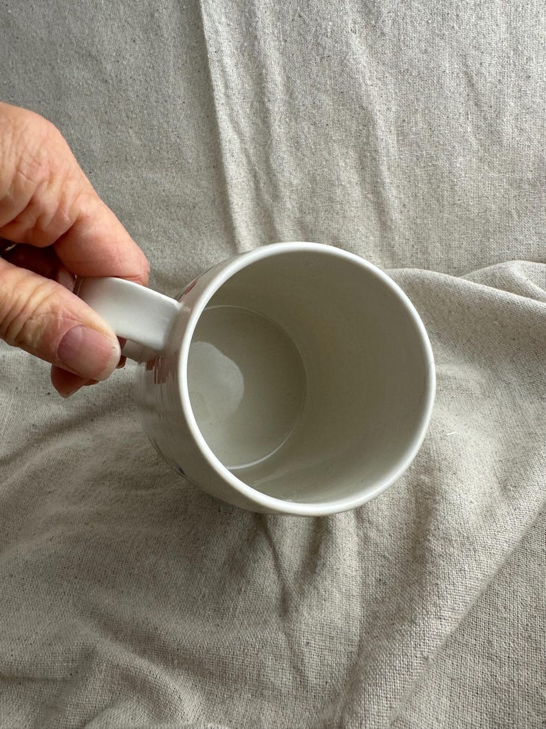 Op de afbeelding: Een witte keramische mok met een gebogen handvat. De mok is leeg en heeft een gladde, glanzende afwerking. De binnenkant van de mok is lichtbeige.