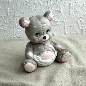 May include: A gray ceramic teddy bear figurine with pink accents on the ears, paws, and belly. The bear has blue eyes, a red mouth, and white polka dots. The bear is sitting on a neutral-colored fabric.