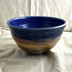 May include: A handmade ceramic bowl with a deep blue interior and a textured exterior. The bowl features bands of color, including shades of blue, brown, and yellow. The bowl is a medium size and appears to be suitable for serving or display.
