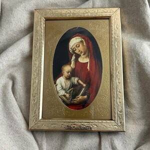 May include: A framed religious artwork featuring a mother and child. The oval image is set within a gold-colored frame with ornate detailing. The mother wears a red robe and white head covering, cradling the child who holds a book.