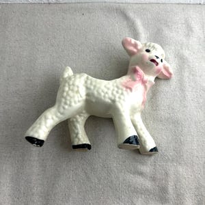May include: A white ceramic lamb figurine with pink ears and a pink bow. The lamb has black hooves and a textured white body.