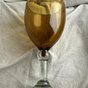 May include: A vintage-style wine glass with a rounded, amber-colored bowl and a clear, slender stem. The base is a dark green color. The glass is likely made of blown glass, showcasing a classic design. The glass is approximately 7 inches tall.