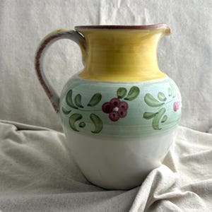 May include: A ceramic pitcher with a white base, a light green band with floral designs, and a yellow top. The handle is brown and white. The pitcher is likely used for serving beverages.