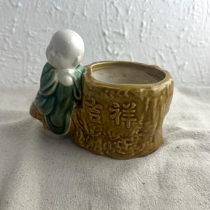 May include: A ceramic planter featuring a small, white-headed figure in a green robe, leaning against a brown, tree-stump-shaped pot. The pot has the Chinese characters for "good fortune" on its side. The planter is suitable for small plants.