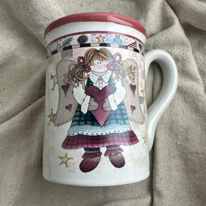 May include: White ceramic mug featuring a whimsical angel design. The angel holds a heart and is adorned with stars and hearts. The rim is red, and the handle is white. A charming and decorative item for beverages.