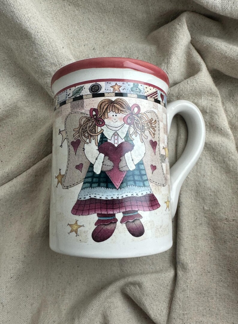 May include: White ceramic mug featuring a whimsical angel design. The angel holds a heart and is adorned with stars and hearts. The rim is red, and the handle is white. A charming and decorative item for beverages.