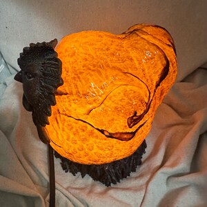 May include: A decorative lamp shaped like a rooster. The lamp has a textured, orange-yellow body with dark brown accents. A detailed, dark brown rooster head is attached to the side. The lamp sits on a dark brown base and emits a warm glow.