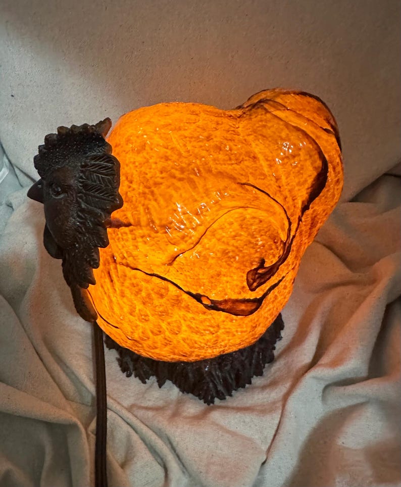 May include: A decorative lamp shaped like a rooster. The lamp has a textured, orange-yellow body with dark brown accents. A detailed, dark brown rooster head is attached to the side. The lamp sits on a dark brown base and emits a warm glow.