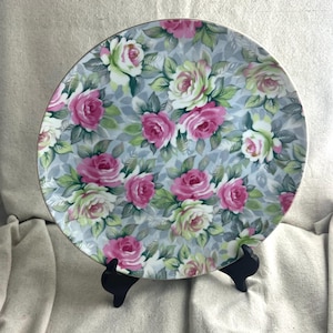 May include: A round decorative plate with a floral pattern of pink, white, and green roses on a light blue background. The plate has a gold rim and is displayed on a black stand.