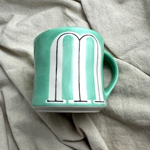 May include: A ceramic mug with a mint green exterior and a white and black letter "M" design. The mug has a rounded shape with a handle on the right side. The mug is sitting on a neutral-colored fabric.