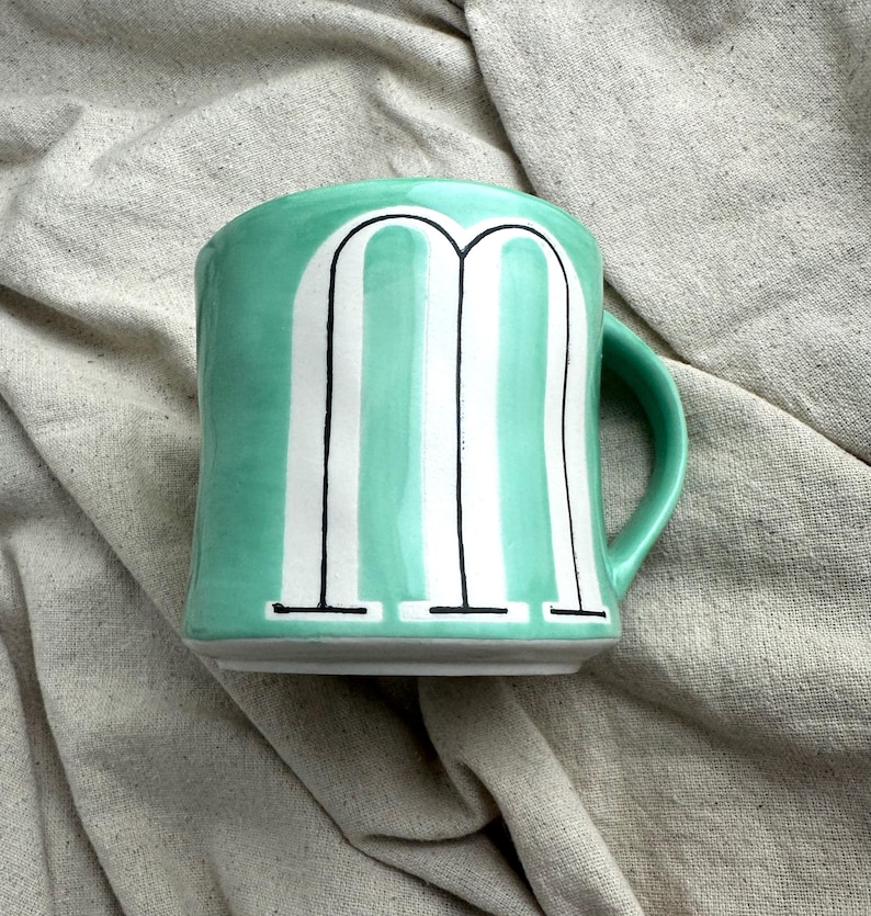 May include: A ceramic mug with a mint green exterior and a white and black letter "M" design. The mug has a rounded shape with a handle on the right side. The mug is sitting on a neutral-colored fabric.