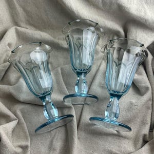 May include: Three light blue, vintage-style sundae glasses with a ruffled rim and a pedestal base. The glasses are made of clear glass and are arranged on a neutral-colored fabric background. The glasses are ideal for serving ice cream.