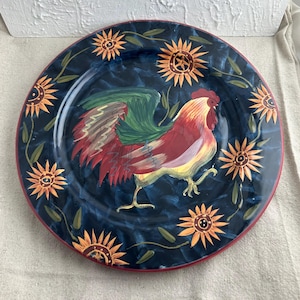 May include: A blue ceramic dinner plate featuring a hand-painted design of a rooster and sunflowers. The rooster is red, yellow, and green, and the sunflowers are yellow and orange.