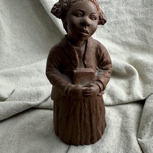 May include: A brown clay sculpture of a young person with textured hair styled in two side pigtails. The figure wears a collared jacket over a skirt and holds a rectangular book with both hands against their chest, featuring a matte finish.