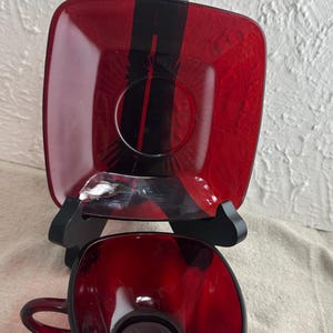 May include: A red glass teacup and saucer set. The square saucer is supported by a black stand. The teacup has a matching red handle. The set is a vibrant red color.