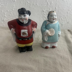 May include: Two porcelain figurines of people wearing traditional Chinese clothing. The male figurine is wearing a red robe with gold accents and a black hat. The female figurine is wearing a blue robe with gold accents and a white hat. Both figurines are standing on a white surface.