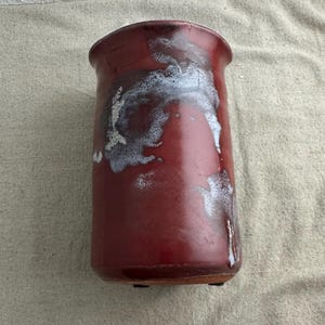 May include: A handmade, cylindrical ceramic utensil holder. The holder is a deep red with white and light grey glaze accents. The piece has a slightly flared rim and sits on a small, flat base. The overall design is rustic and functional.