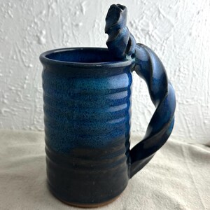 May include: A handmade ceramic mug with a unique twisted handle. The mug features a gradient of blue hues, transitioning from a deep navy at the base to a lighter blue at the top. The mug is cylindrical with a textured surface.