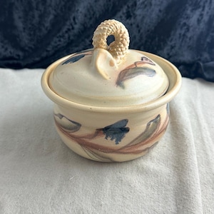 May include: A small, cream-colored ceramic jar with a lid. The jar has a floral design painted on it in shades of brown, blue, and green. The lid has a unique, textured knob.