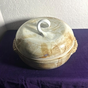 May include: A round, lidded ceramic serving dish with a white, curved handle. The dish has a beige and brown marbled pattern and a rope-like design around the rim. The dish is sitting on a purple surface.