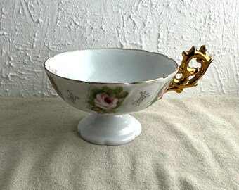May include: White teacup with a pedestal base and a gold handle. The cup features a floral design with pink roses and green leaves. The rim and handle are trimmed with gold. A decorative piece for serving tea.