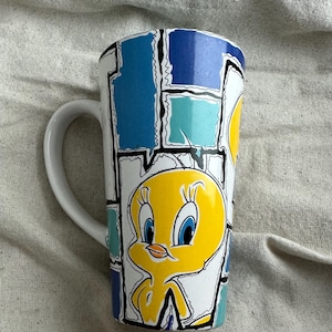 Peut inclure: Mug en céramique blanche de grande taille avec une image de dessin animé de Titi. Le mug présente un motif répétitif de formes géométriques bleues, turquoise et blanches. La poignée est blanche.